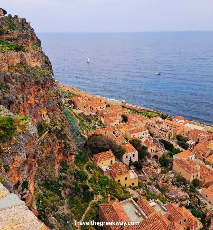 Aerial view of the medieval walled town of Monemvasia, Greece, showing red-tiled roofs nestled between a steep cliff and the blue Aegean Sea.