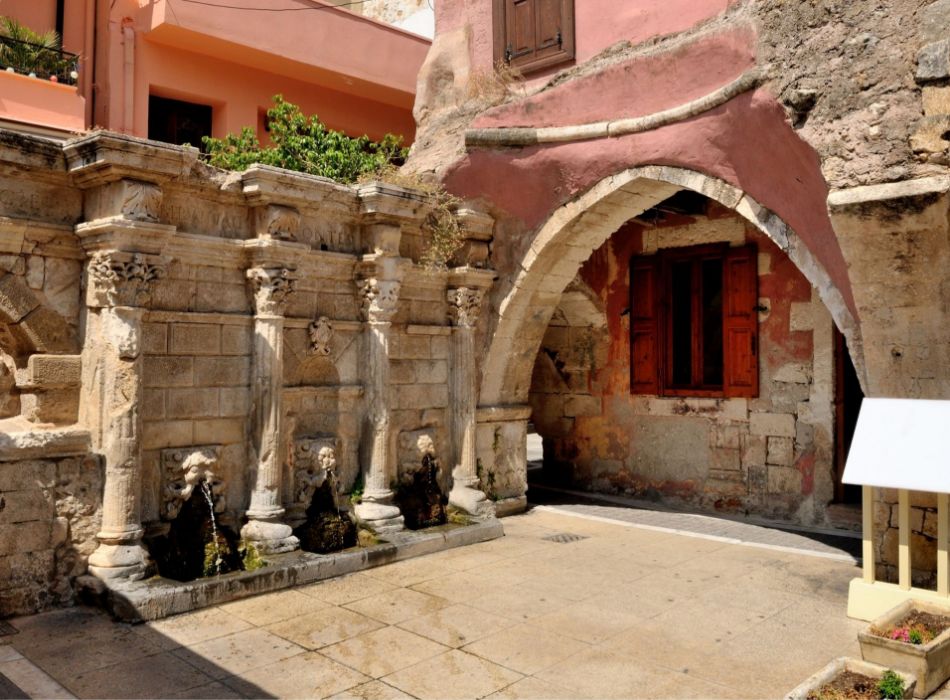 Historical Venetian Rimondi Fountain in Rethymno, Crete, featuring stone lion heads and arched water spouts set against a background of pink-washed buildings.