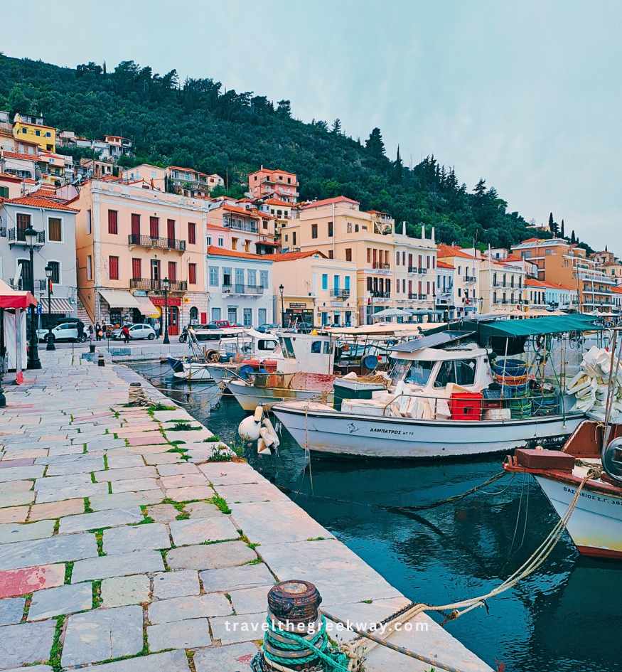 The charming harbor of Gythio in the Peloponnese, Greece with several small fishing boats moored along a stone pier, backed by multi-story colorful buildings at the foot of a lush, green hill.