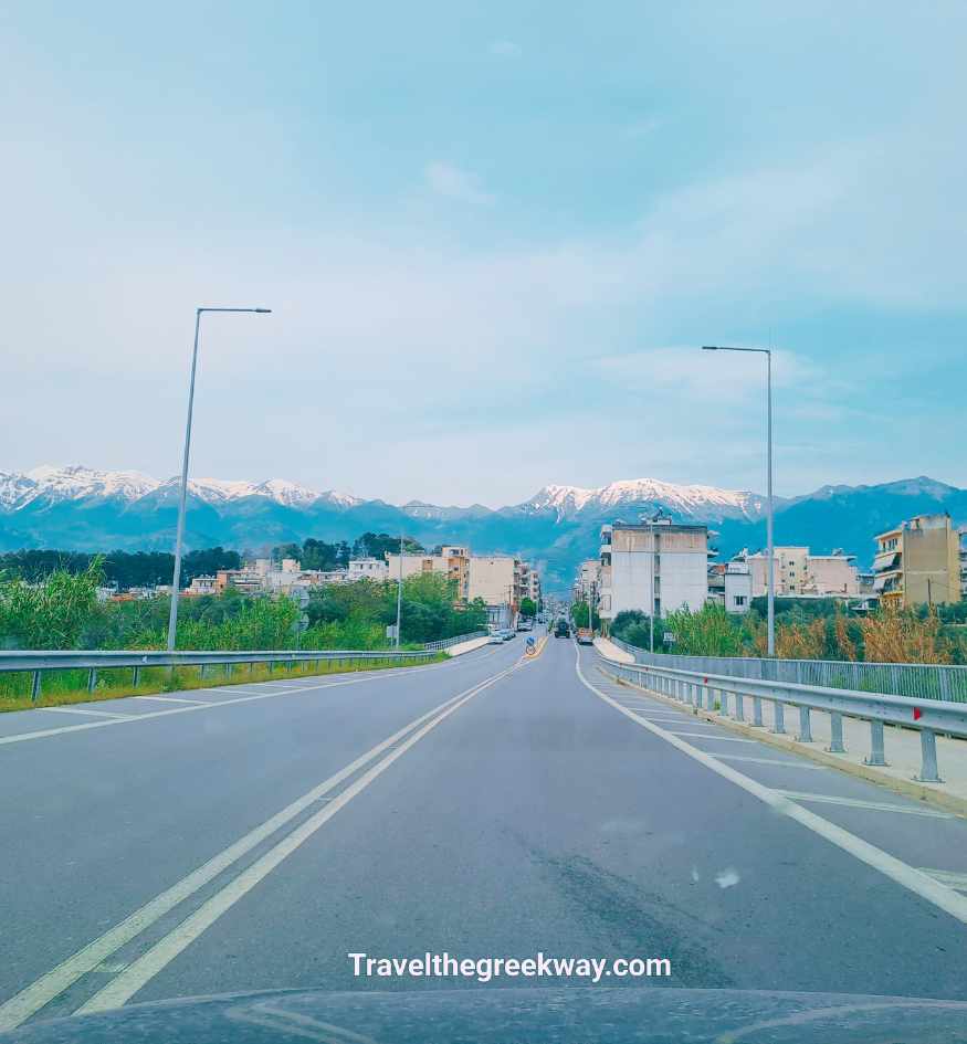View from a car looking down a straight highway toward Sparta in Greece, with a stunning backdrop of high, snow-capped mountains under a pale blue sky.