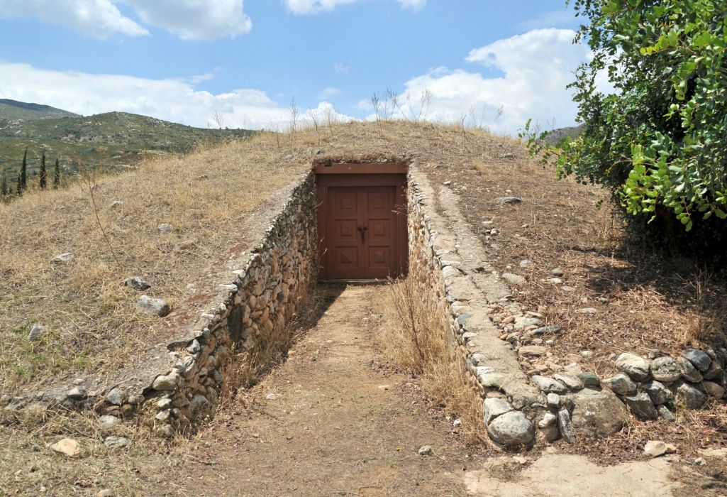 Marathon Timuli Tomb of Plataea.An ancient burial mound in Marathon, Greece, featuring a stone-lined entrance path leading to a wooden door, set against a backdrop of rolling hills and a partly cloudy sky.