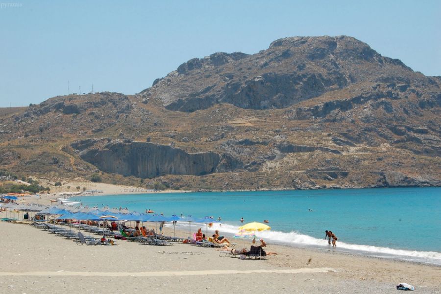 Sandy beach in Plakias, Rethymno, Crete, with sunbathers under umbrellas and a backdrop of rocky hills.