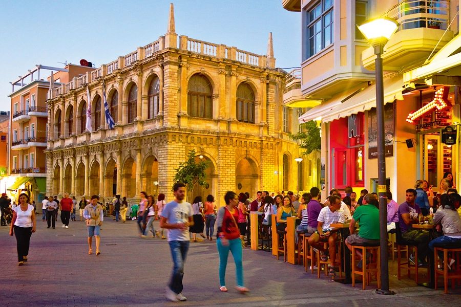 Vibrant street scene in Heraklion, Crete, Greece, with people enjoying an evening out at cafes and restaurants near a historic building under a twilight sky.