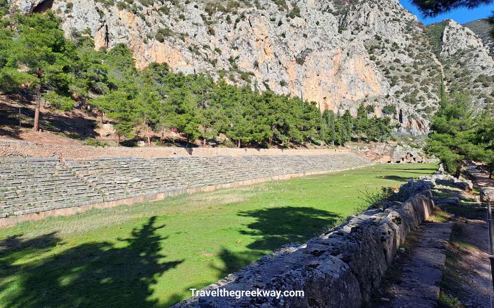 The ancient Stadium of Delphi set beneath towering cliffs and surrounded by pine trees.
