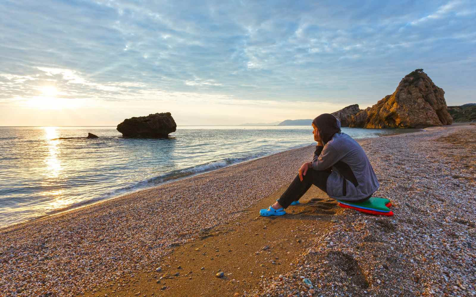 Woman sitting on Potistika Beach in Pelion, Greece, admiring the sunset over the Aegean Sea with rocky formations in view.