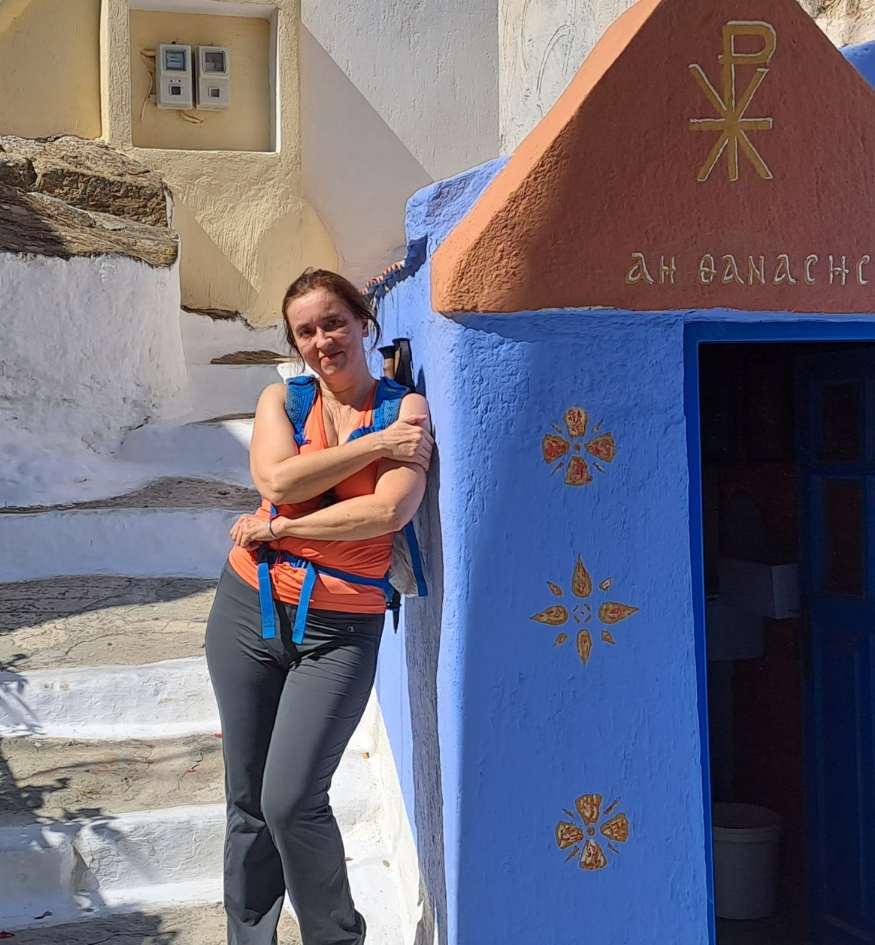 Evgenia of Travel the Greek Way leaning against a vibrant blue and terracotta-roofed traditional chapel in Ioulida, Kea island, Cyclades, Greece.