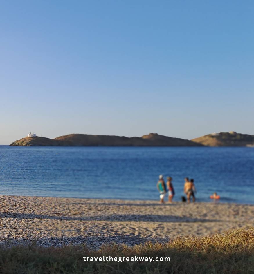 People in the distance on a pebbled beach looking out at the sea, with a small white lighthouse visible on a point in the distance under a clear sky, in Kea island Greece.