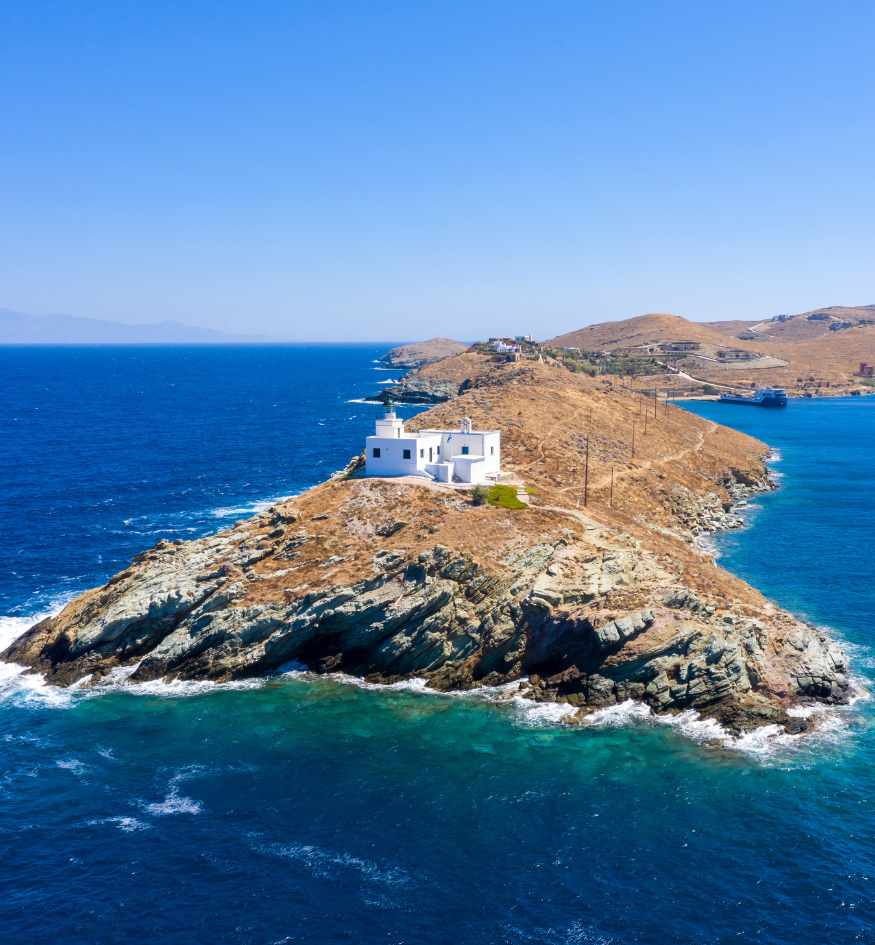 Elevated view looking down a narrow point of rocky land to a small, isolated white lighthouse building surrounded by the blue sea, in Kea island Greece.