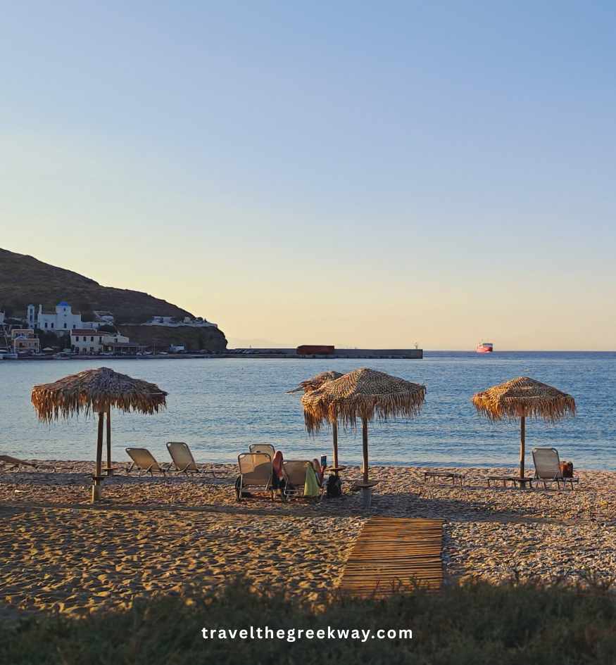 A pebbled beach with straw umbrellas and sunbeds at the water's edge, against a backdrop of a small coastal village and hills at sunset in Kea island, Greece.