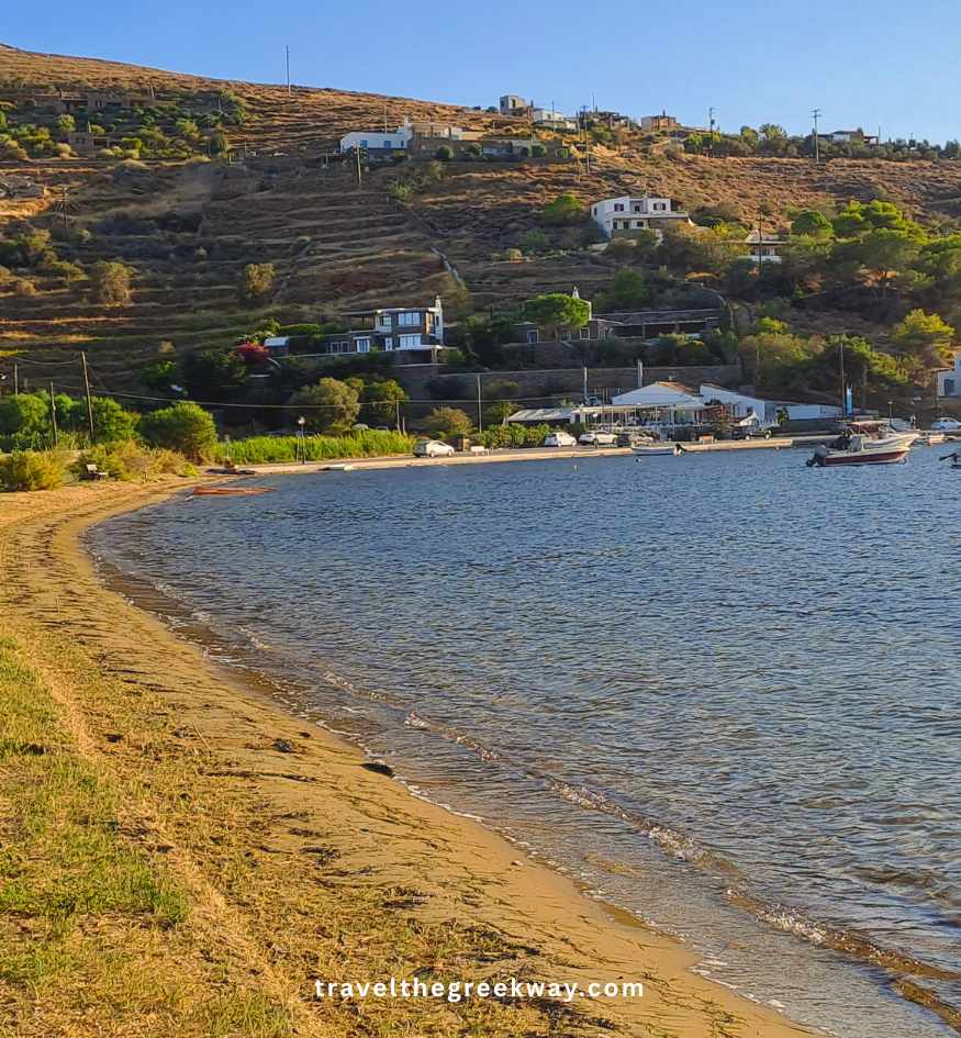 Gentle waves on a sandy beach under a bright blue sky, with modern waterfront buildings and a tree-lined hillside in the background, in Kea island Greece.