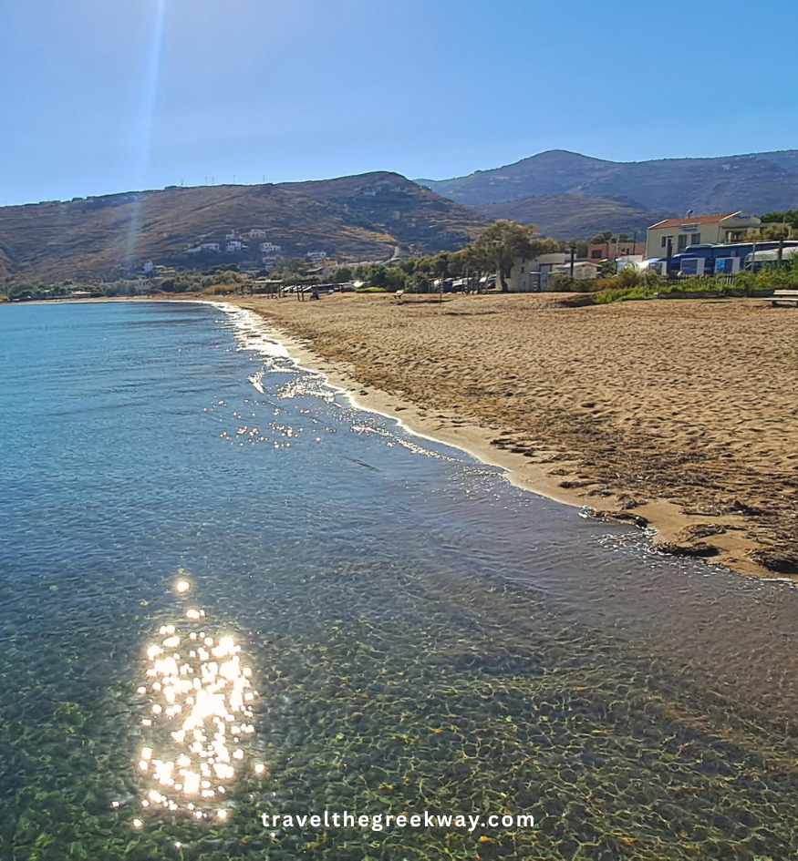 Gentle waves on a sandy beach under a bright blue sky, with modern waterfront buildings and a tree-lined hillside in the background, in Kea island Greece.
