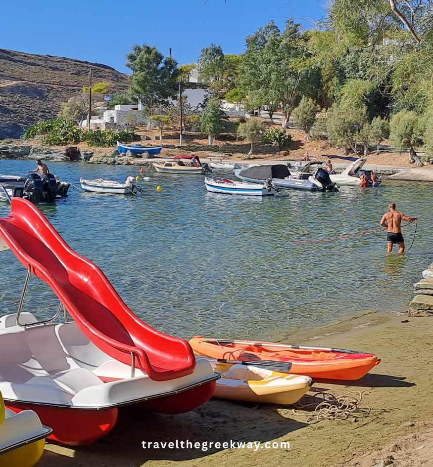 A pebbled beach with a red slide pedal boat, a kayak, and various motorboats moored in a calm cove, with people in the water and a tree-lined shore, in Kea island Greece.