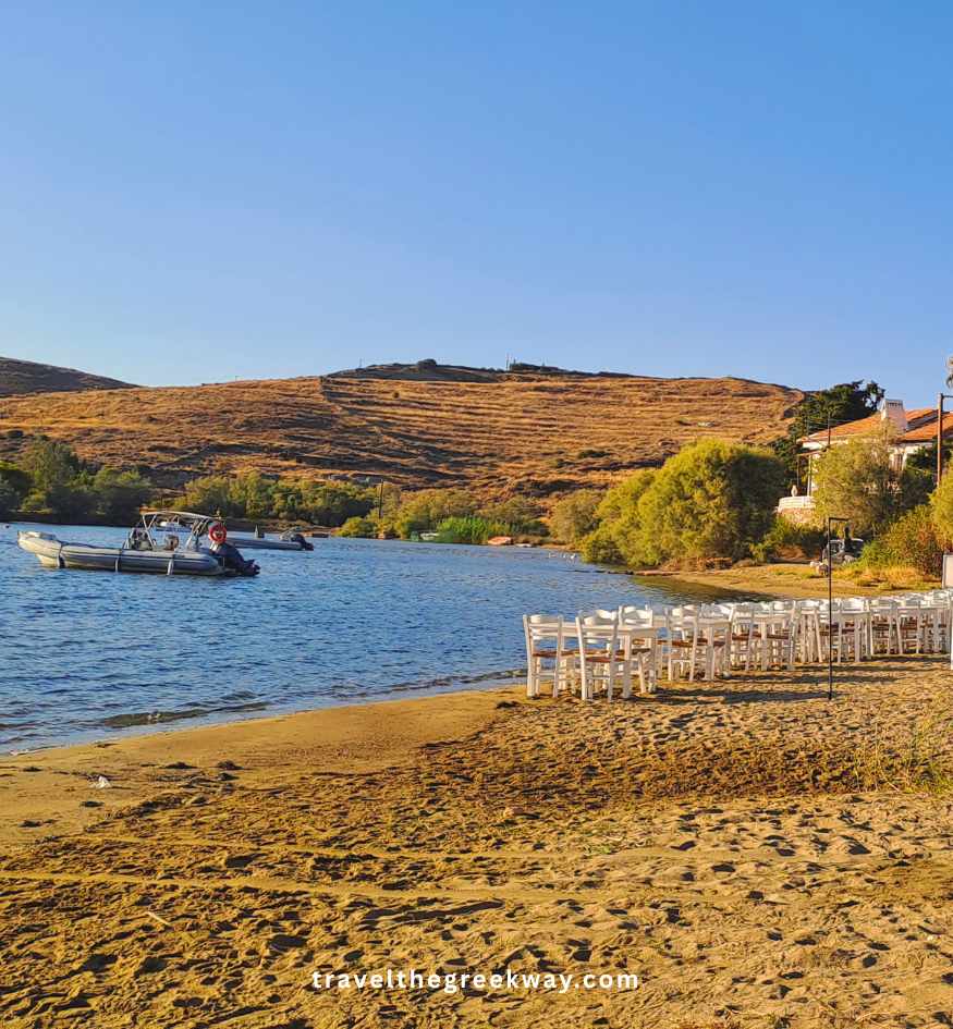Rows of white wooden chairs with cane seats on a sandy beach at sunset, with a motorboat on the water, trees, and hills, in Kea island Greece.