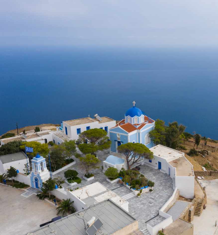High-angle view of a serene blue-domed church and surrounding white buildings on a coastal cliff, against the vast expanse of the open blue Aegean in Kea Island.