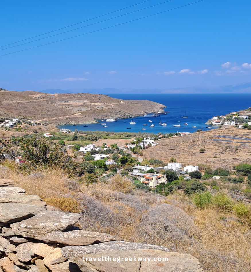 A panoramic view over an arid landscape to a large, calm blue bay with numerous yachts and sailboats anchored off a small village, on Kea Island.