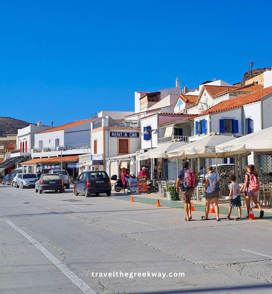 Tourists walking along the sunny waterfront street lined with shops and car rentals in Korissia, Kea, Cyclades, Greece.