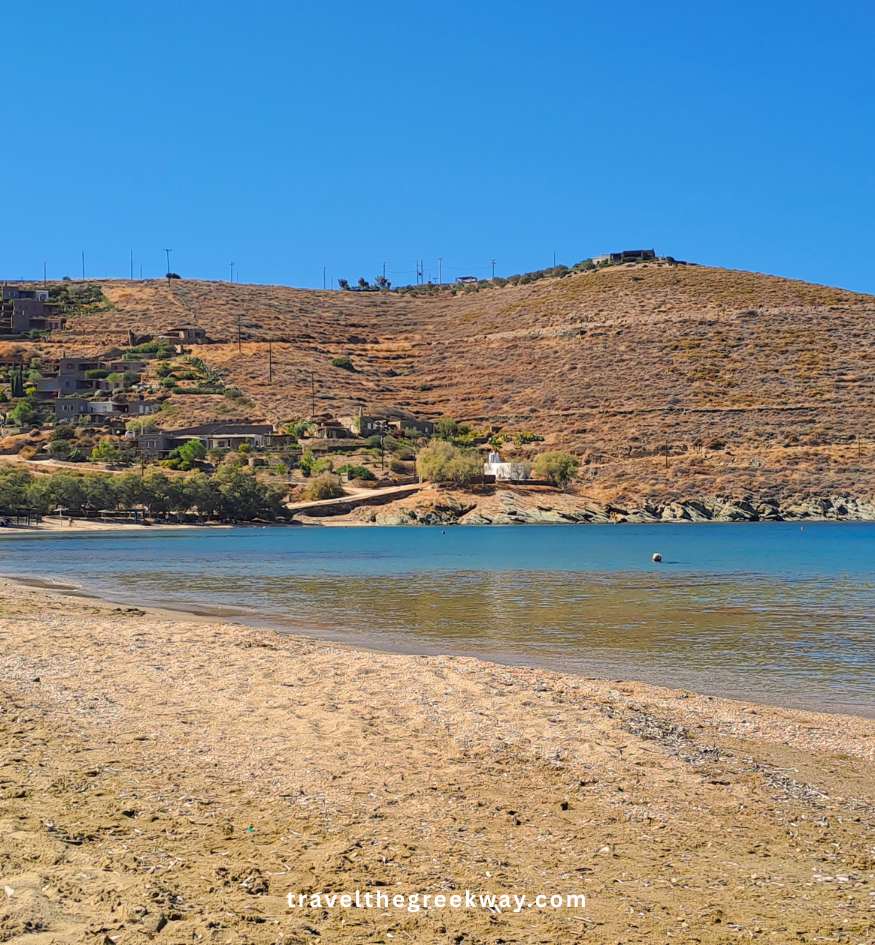 A wide view of a quiet, golden sandy beach with blue water and arid hills in the background on Kea island, Cyclades, Greece.
