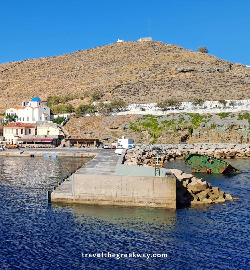 A green shipwreck resting in the blue water near the stone pier and white buildings of Kea island, Cyclades, Greece.