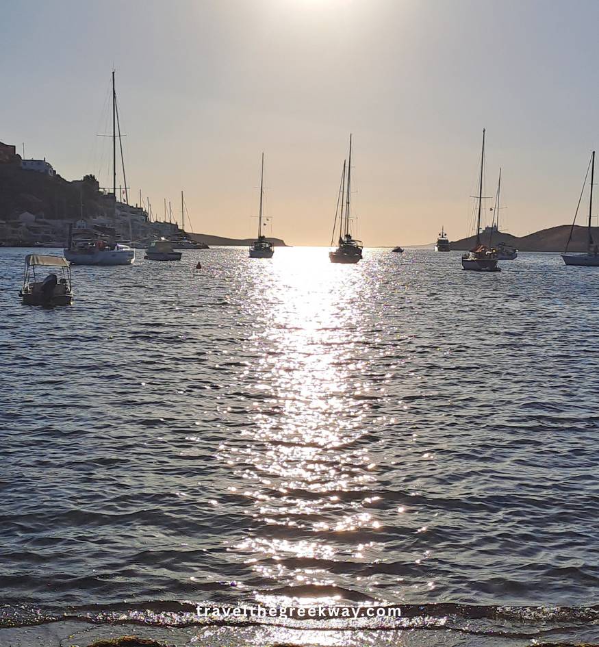 A panoramic view over an arid landscape to a large, calm blue bay with numerous yachts and sailboats anchored off a small village, in Kea island Greece.