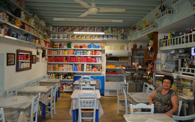 The image depicts the interior of a quaint Greek taverna on Folegandros Island. The space is cozy, with a rustic charm, featuring a simple decor of wooden chairs and tables covered in plastic tablecloths. Shelves along the walls are stocked with an array of colorful packaged goods, adding a vibrant backdrop. Fresh produce is displayed on the counter, and a refrigerator in the corner holds beverages. The ceiling fan and light blue-painted ceiling beams add to the inviting atmosphere. A woman, presumably the proprietor, sits at one of the tables, adding a personal touch to this authentic local dining experience.