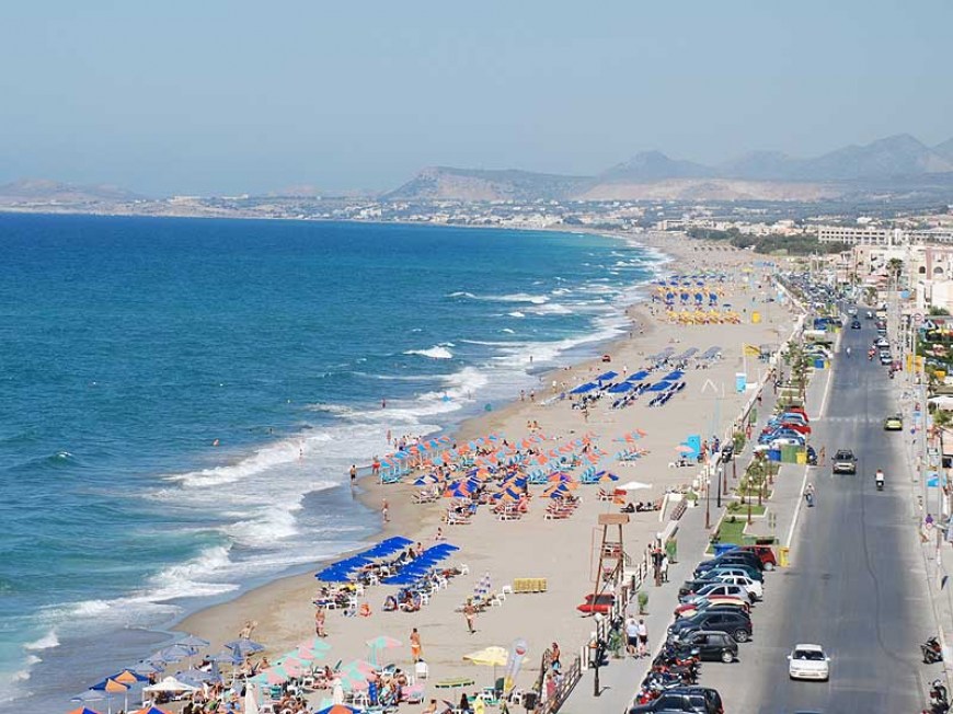 Long stretch of sandy beach in Rethymno, Crete, with rows of colorful umbrellas and sunbathers, adjacent to a busy coastal road lined with parked cars.