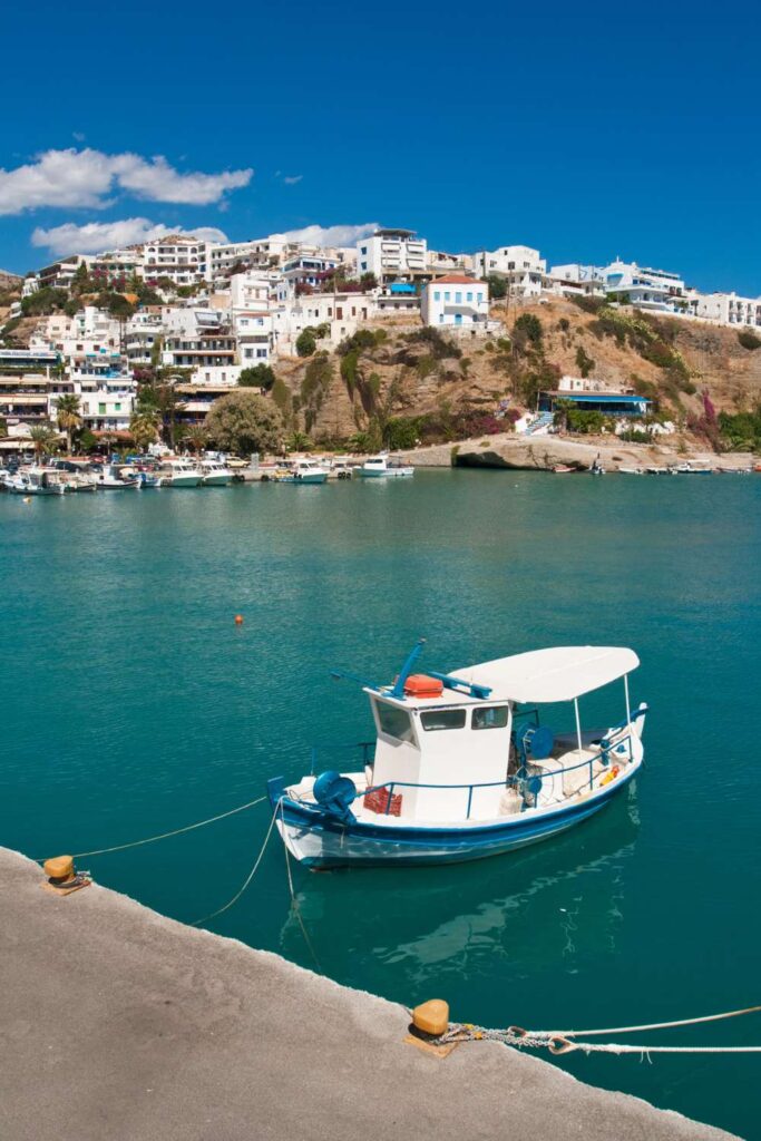 Scenic view of Agia Galini in Rethymno, Crete, featuring a traditional fishing boat docked in the calm blue waters, with a hillside town of white-washed buildings in the background.