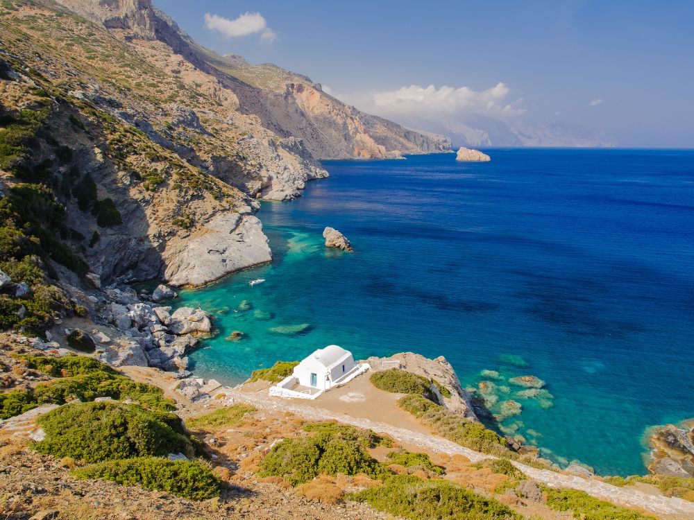 Breathtaking view of a small white chapel by the turquoise waters of a secluded bay on Amorgos Island, Greece, surrounded by rugged cliffs and lush greenery under a clear blue sky.