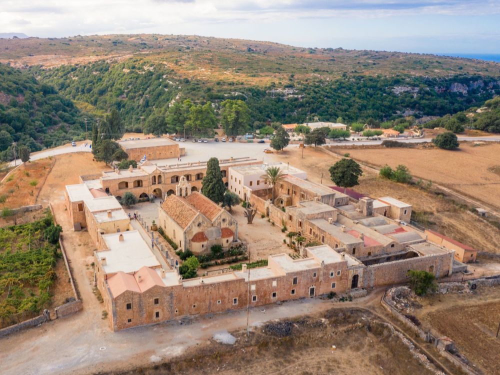 Aerial view of Arkadi Monastery, Rethymno, Crete, showcasing its historic stone buildings and red-tiled roofs, set in a lush, hilly landscape.