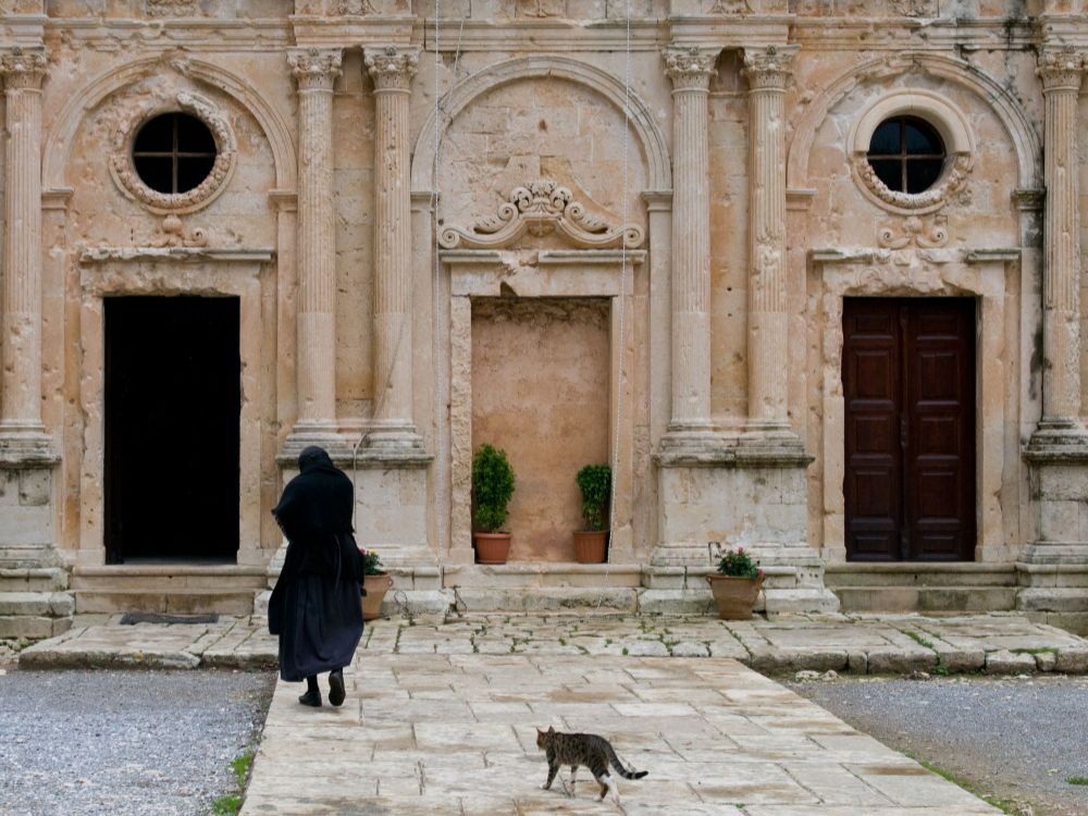 Facade of Arkadi Monastery in Rethymno, Crete, featuring intricate stonework and arched doorways, with an elderly woman and a cat walking in front.