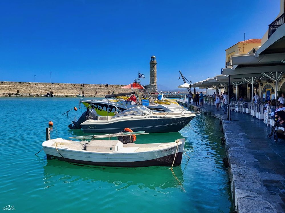 Picturesque harbor of Rethymno, Crete, with boats docked along the waterfront, a stone lighthouse in the distance, and cafes and restaurants lining the promenade.