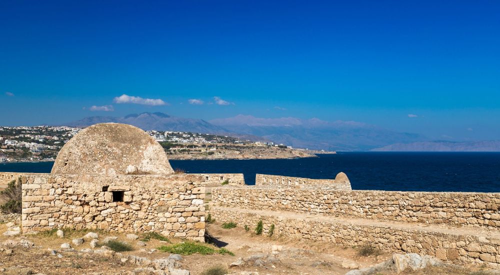 Ancient stone fortifications of the Fortezza of Rethymno, Crete, with a dome-shaped structure and panoramic views of the coastline and distant mountains.