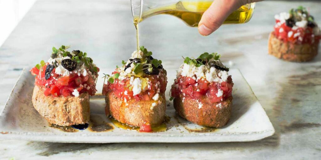 Delicious Dakos dish from Crete, featuring rusks topped with chopped tomatoes, feta cheese, olives, and herbs, being drizzled with olive oil.