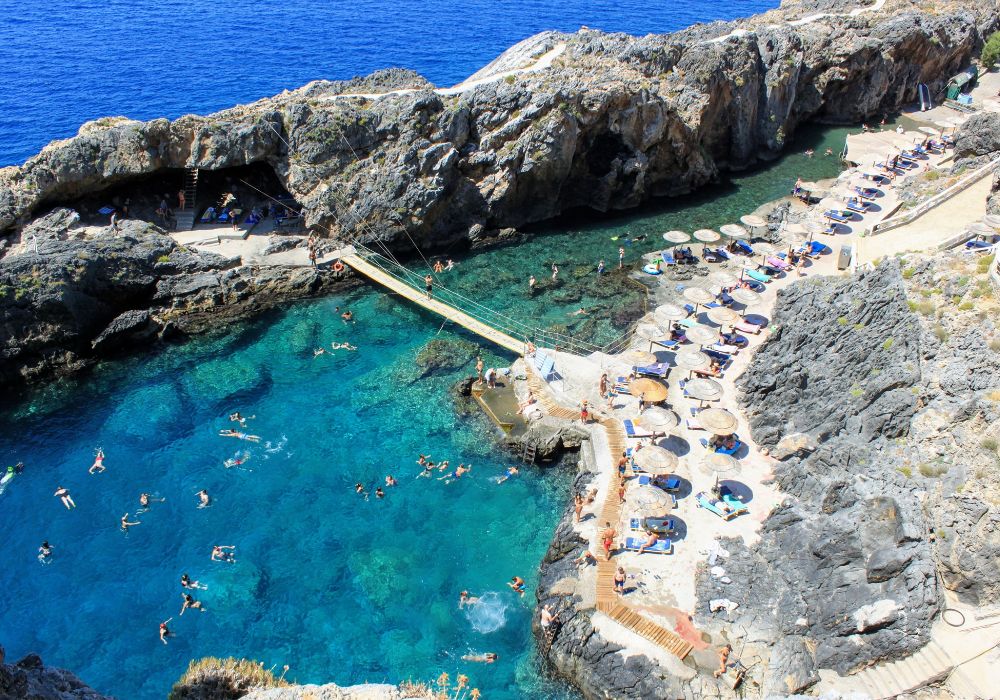 Scenic view of Kalypso Beach in Rethymno, Crete, with crystal-clear turquoise waters, rocky cliffs, and sunbathers relaxing on sun loungers.