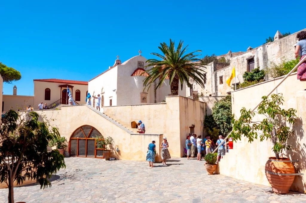 Visitors exploring the courtyard of Preveli Monastery in South Crete, Greece, with white-washed buildings and palm trees under a bright blue sky.