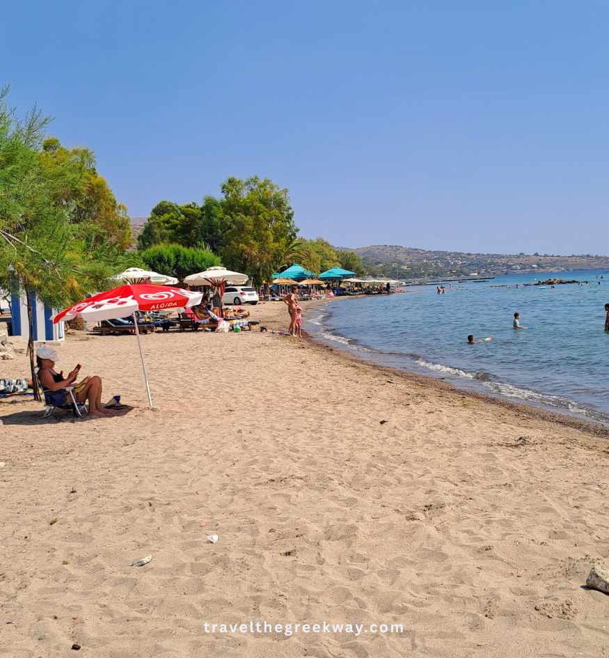 Sunny Marathonas A beach on Aegina Island, Saronic Greece, featuring a wide sandy shore, beach umbrellas, and tourists swimming in the sea.