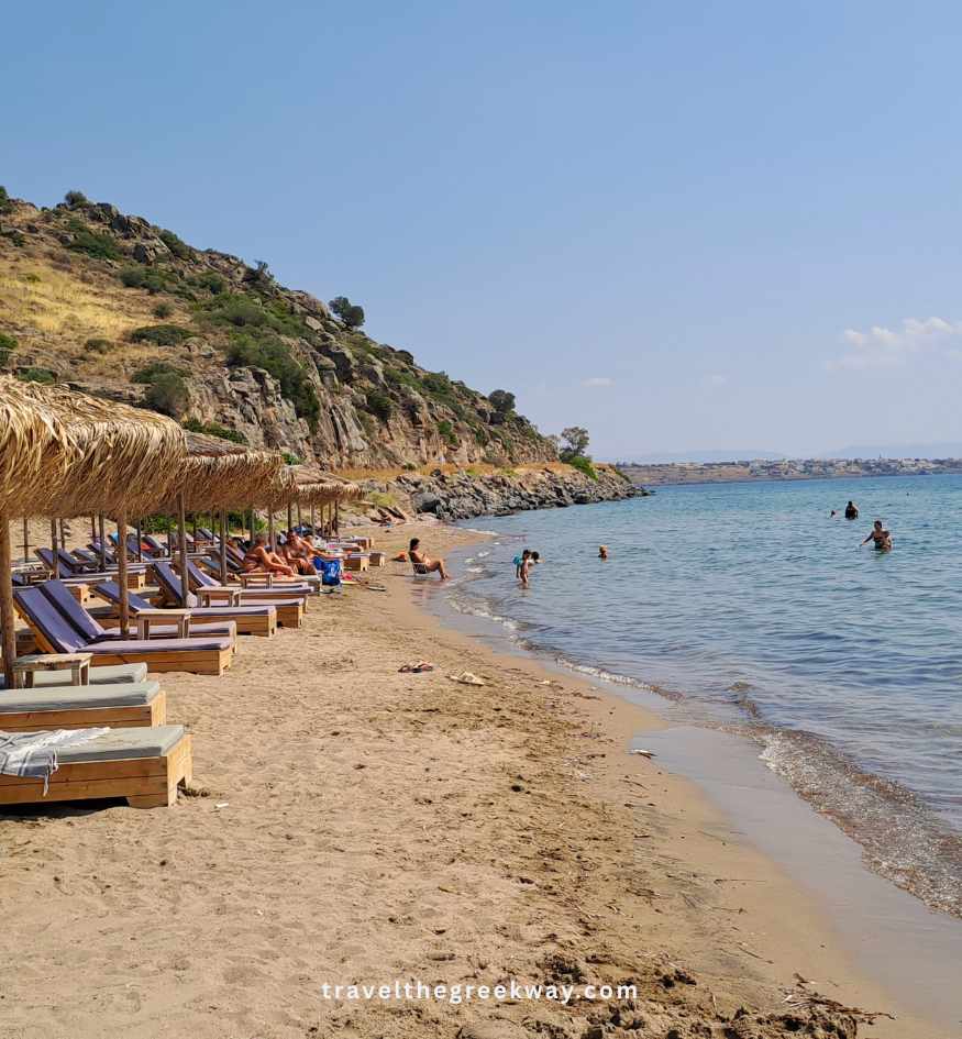 Row of straw umbrellas and sunbeds on the sandy shoreline of Marathonas Beach, Aegina Island, Saronic Greece, under a clear blue sky.