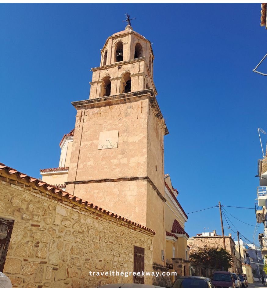 A tall, historic stone church bell tower stretches into a clear blue sky on Aegina island, Saronic, Greece.
