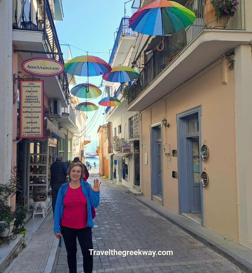 Evgenia of Travel the Greek Way waving in a narrow street of Aegina Town, Aegina Island, Saronic Greece, decorated with colorful hanging rain umbrellas overhead. Where to stay in Aegina