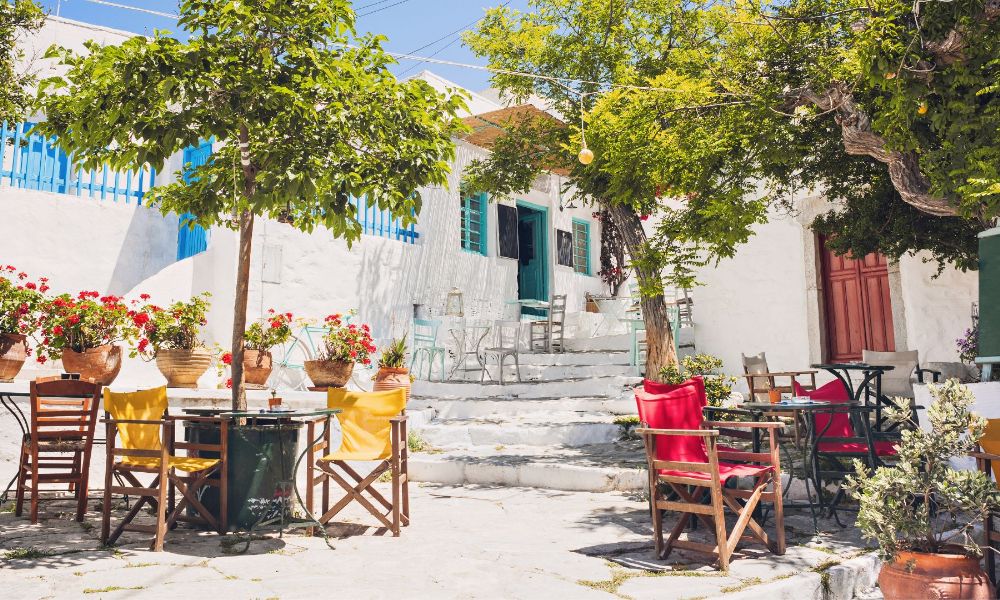 A square with tables and chairs and trees and whitewashed steps in Amorgos Island Greece.