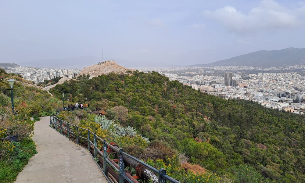 A panoramic view from Lycabettus Hill in Athens, overlooking the city’s green spaces and distant mountains under a cloudy sky.