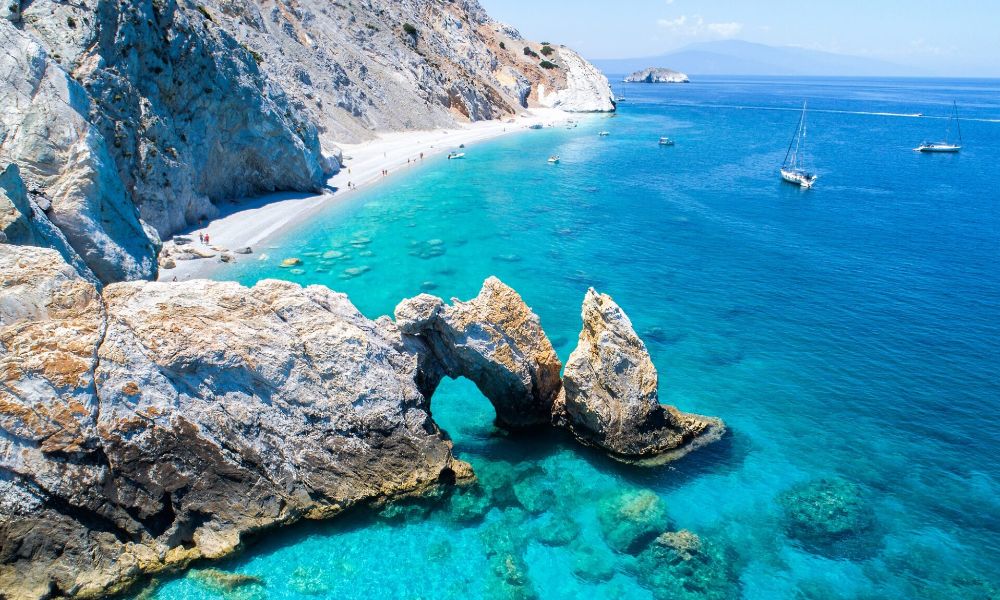 A captivating sight of a sandy coastline flanked by verdant pine trees, where the shimmering turquoise waters of Skiathos' beaches merge seamlessly with the horizon, creating a breathtaking vista.