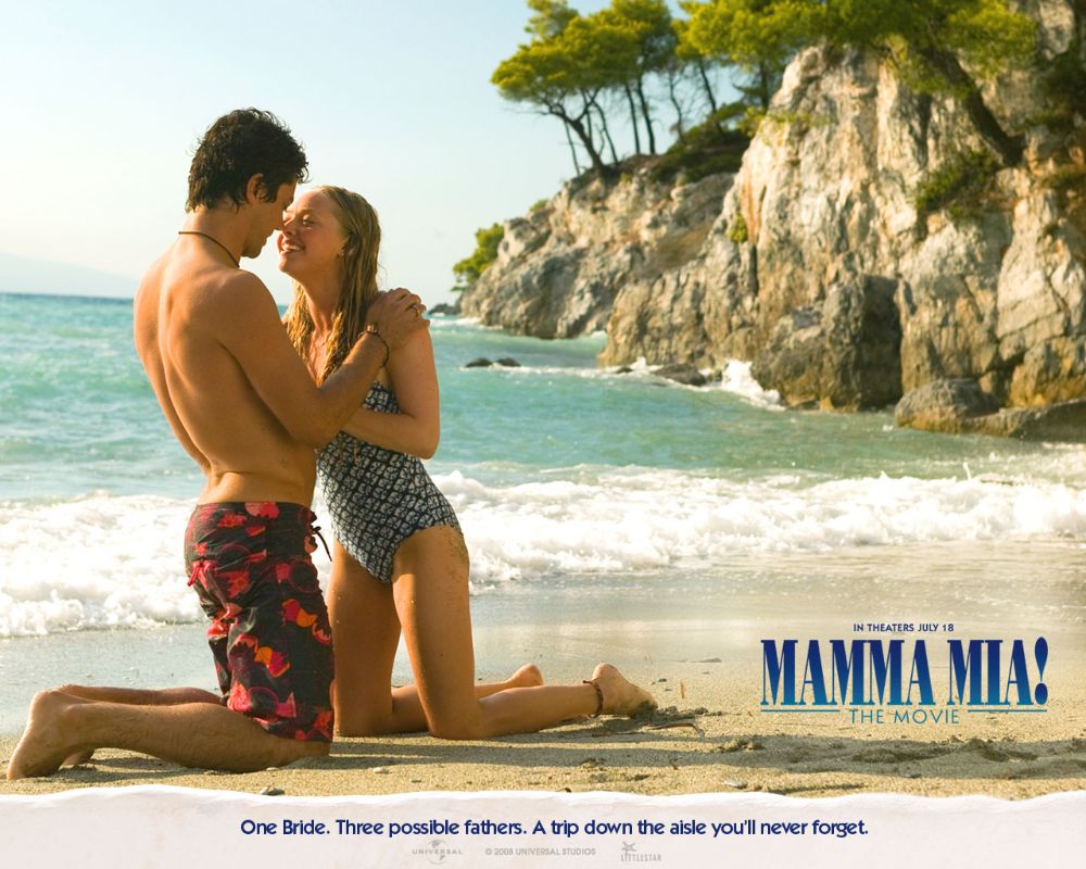 A couple kneels on a sunny beach, smiling at each other with clear blue waters and rocky cliffs in the background. It is from the Mamma Mia film.