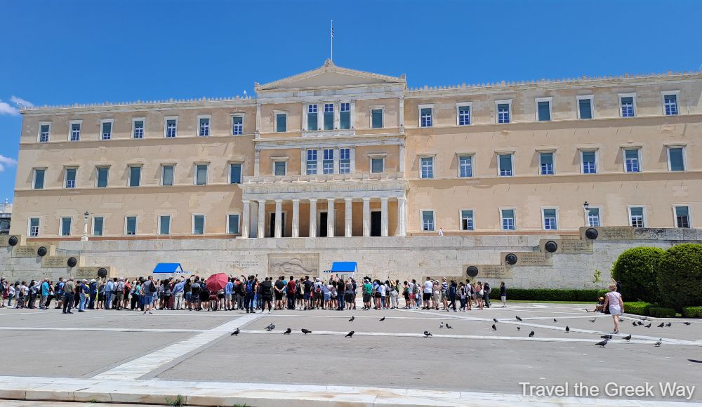 A wide view of the Hellenic Parliament in Syntagma Square, Athens, with crowds gathered in front of the building.