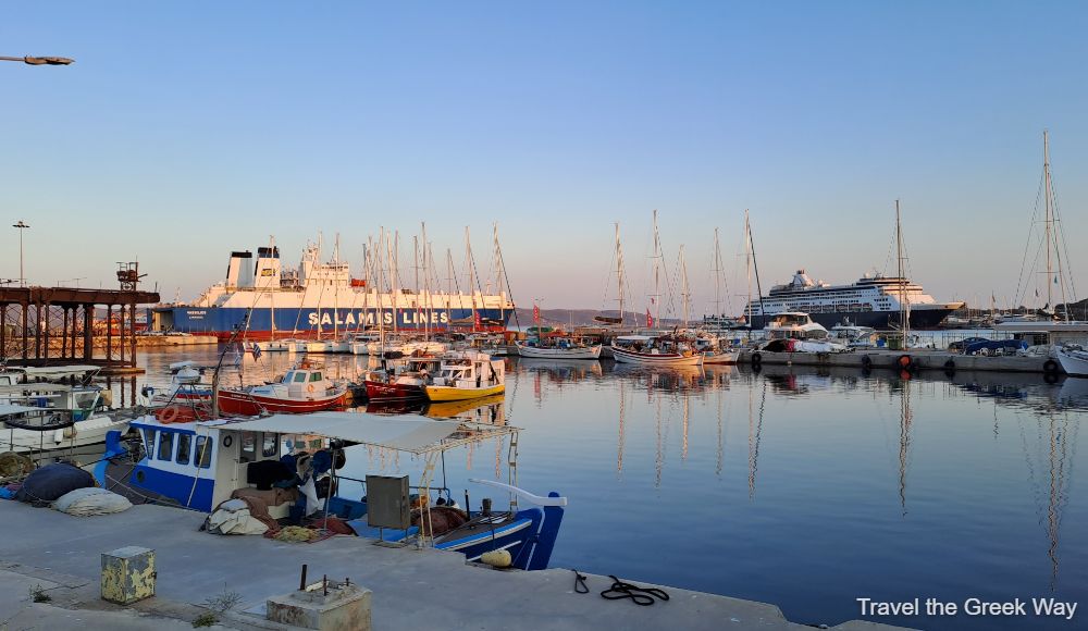 View of Lavrio Port in Greece, featuring various boats docked along the waterfront. The scene includes colorful fishing boats in the foreground, larger yachts and sailboats in the middle, and a large ferry and cruise ship in the background. The calm water reflects the vessels and the clear sky at sunset, creating a serene harbor atmosphere. The distant hills add depth to the port landscape.