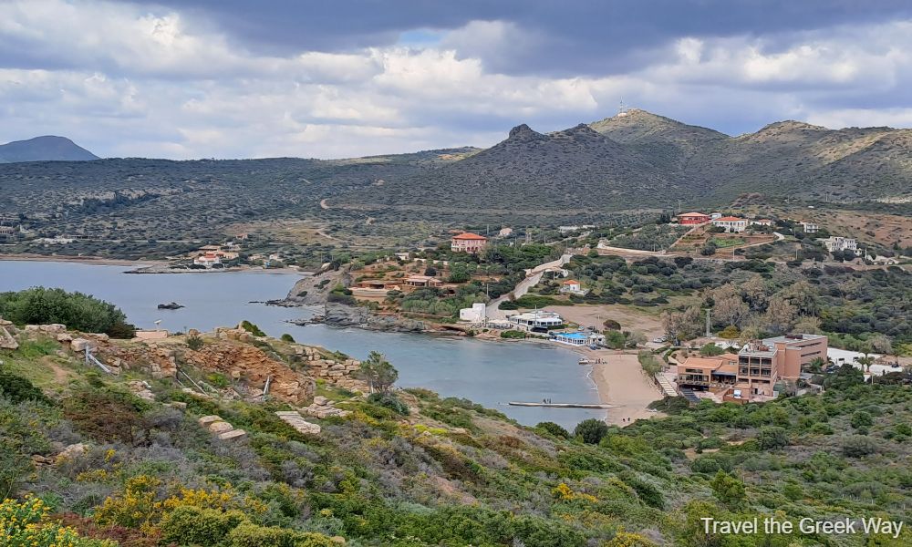 View of a coastal village near Cape Sounion, Greece, with a sandy beach and a small bay surrounded by hills. The scene features scattered buildings and homes nestled among lush green vegetation. A winding road connects the village to the surrounding landscape, and the sea is calm under a partly cloudy sky. In the foreground, ancient stone ruins are visible, blending history with the natural beauty of the area.