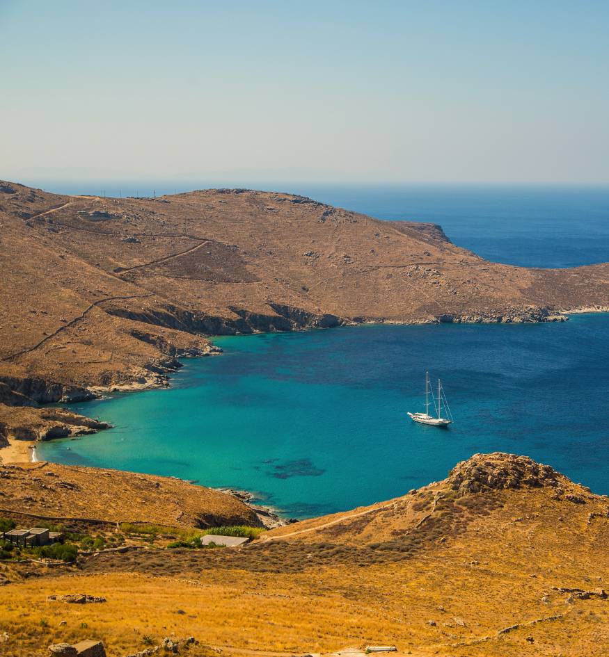 A luxury yacht anchored in a secluded, crystal-clear turquoise bay surrounded by the dry, golden hills of Serifos, Greece.