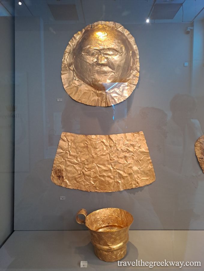 Golden funeral mask and decorative shield and cup from Mycenae, Greece, displayed in a museum.