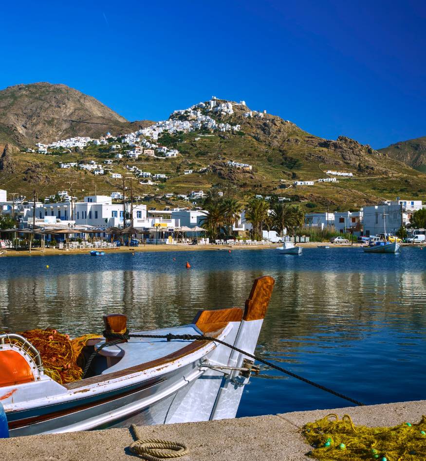 A traditional fishing boat in the calm blue waters of Livadi port with the Chora village on the hill behind, Serifos, Greece.