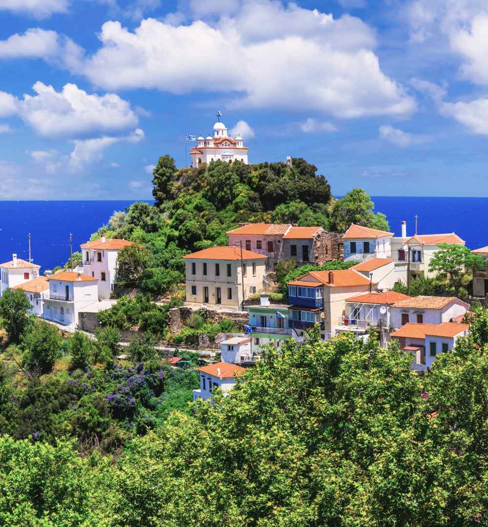 A hilltop white church with a red dome overlooks traditional orange-roofed houses in Karlovasi, Samos, Greece.