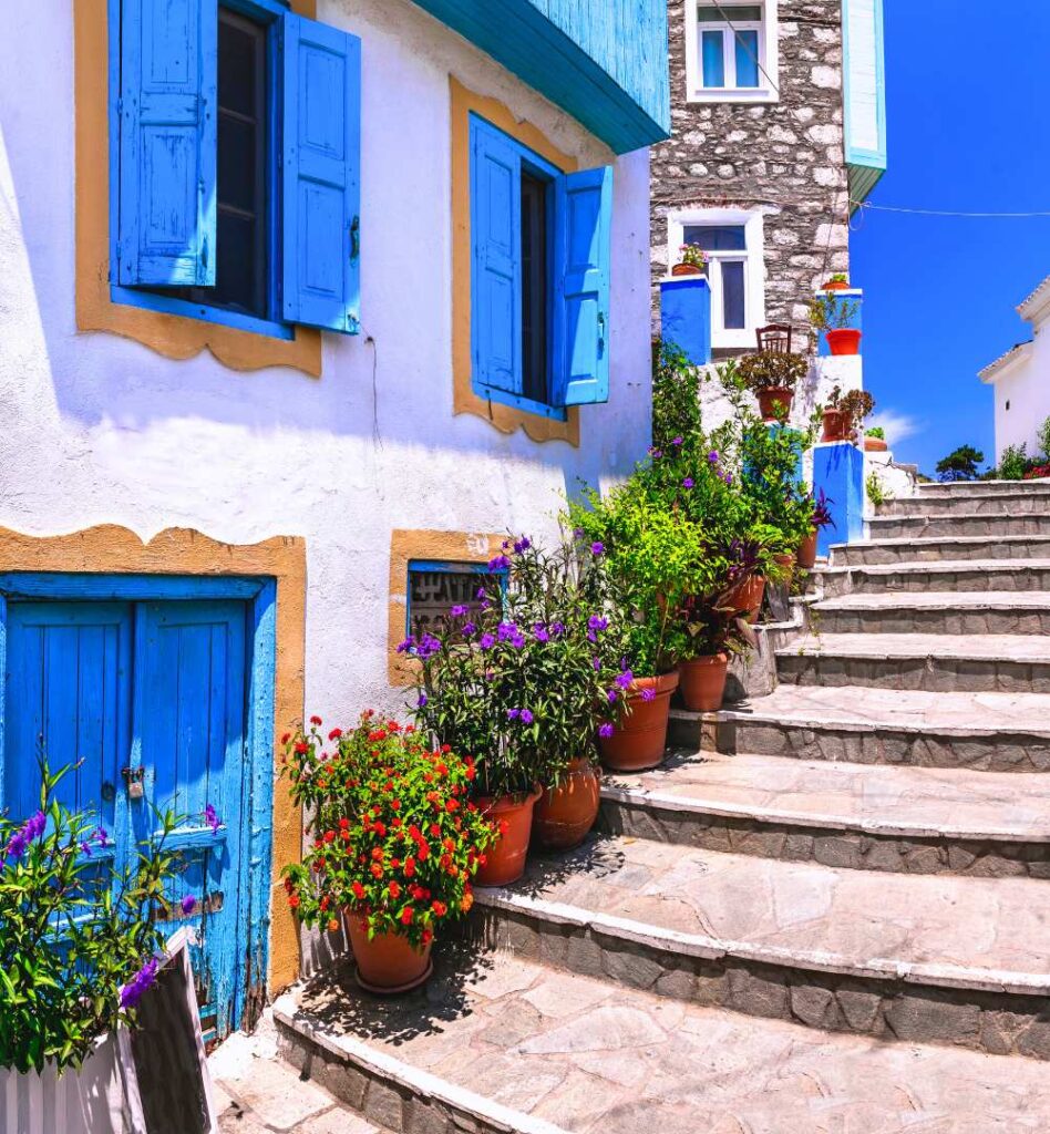 Narrow stone staircase in Kokkari, Samos, Greece, lined with flowers and white houses with bright blue shutters.